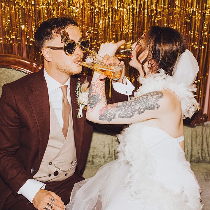 Bride & groom sit in DIY photo booth wearing fun sunglasses and gold streamers to the back