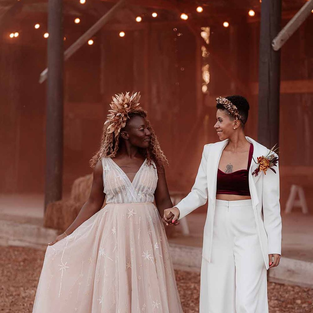 Bride in elegant white silk suit with autumnal dried floral boutonniere holding hands with bride in sleeveless floaty peach gown with embroidered stairs and gold bridal crown 