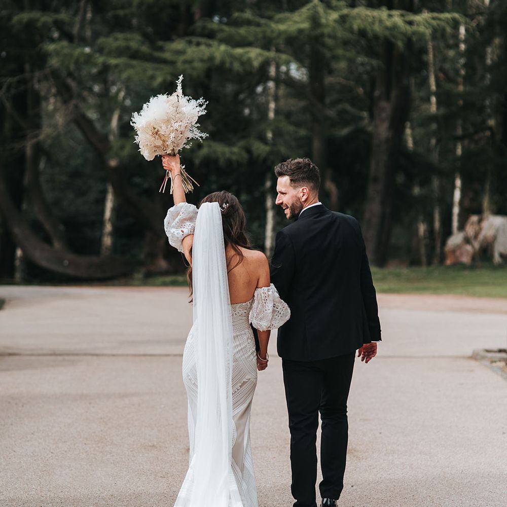 Bride in boho lace wedding dress with train raises her white dried flower bouquet as she walks away with the groom in a black suit 