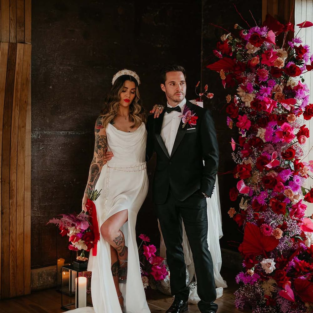 Bride in cowl neck sleeveless satin wedding dress with leg slit, gold glittery wedding headband and groom in classic black tuxedo with magenta amaryllis and orchid boutonniere posing by pillar candles in large glass black metal lanterns and large floral decorations with hot pink, bright pink and magenta bridal bouquet with dahlias, orchids, roses, amaryllis, anthuriums and dried flowers 