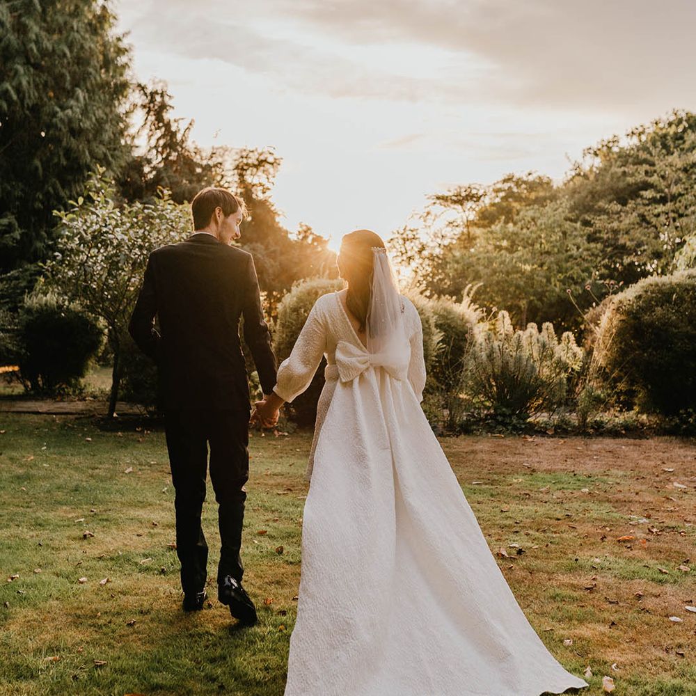 The groom in black tie walks hand in hand with the bride wearing a short wedding dress with a train and big bow detail during sunset 