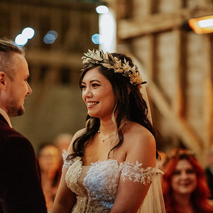 The bride in a delicate lace off the shoulder wedding dress with boho dried flower crown stares into the groom's eyes for their ceremony