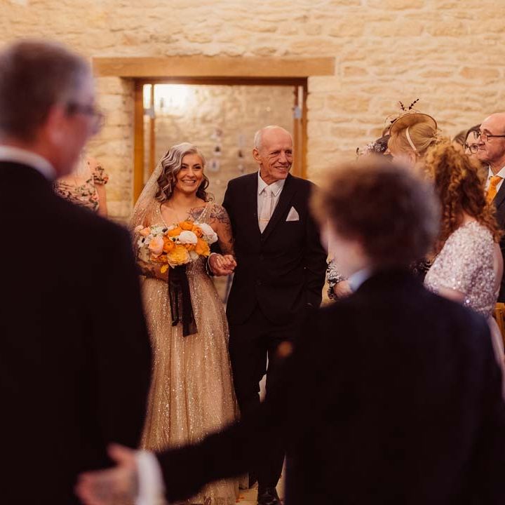 Bride in sleeveless deep v neck sparkly gold wedding dress with sheer and gold polka dot wedding veil holding orange garden rose, white poppy, light pink carnation and foliage bridal bouquet wrapped in sparkly gold ribbon with father of the bride walking down the aisle at Kingscote Barn Tetbury wedding venue