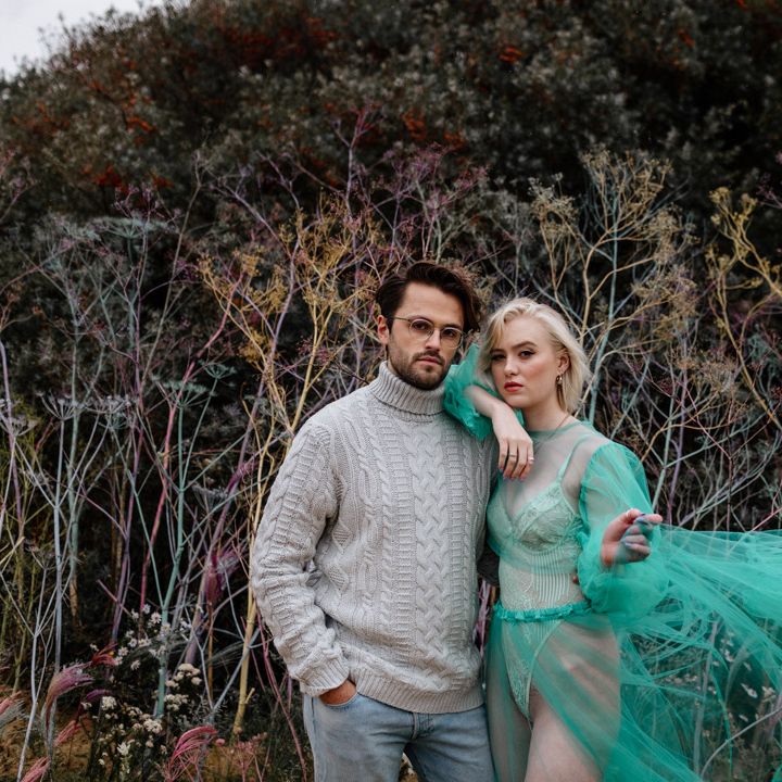 Brunette bearded groom in glasses, sweater and jeans with his bride in a sheer tulle wedding dress on the beach 