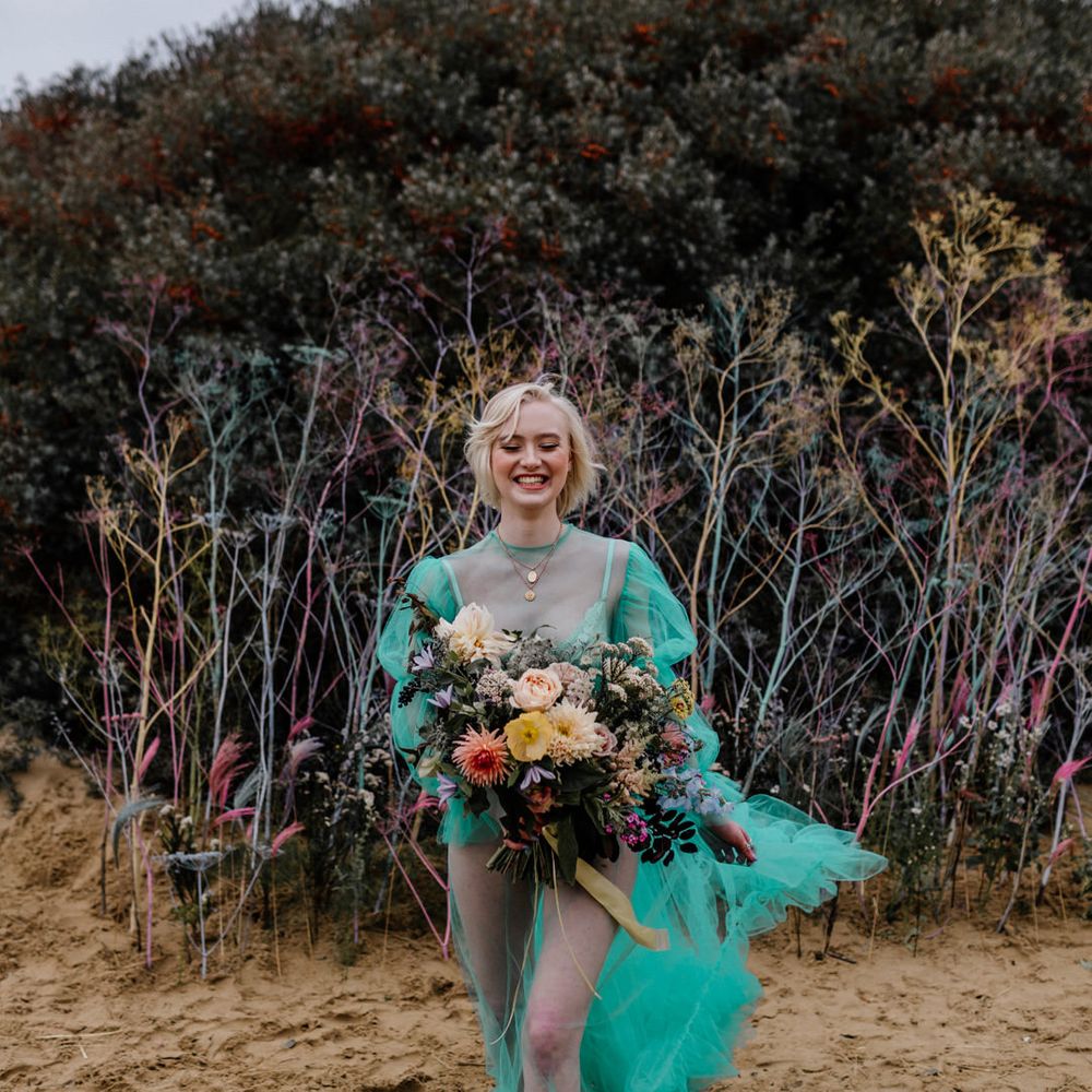 Barefoot bride walking in the sand at her beach elopement in a pastel wedding dress holding a colourful flower wedding bouquet tied with ribbon