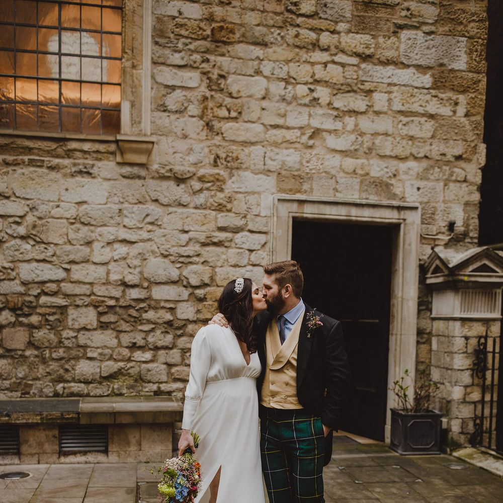 Bride and groom kissing. Bride wears long sleeved wedding dress with front split and groom wears Trews