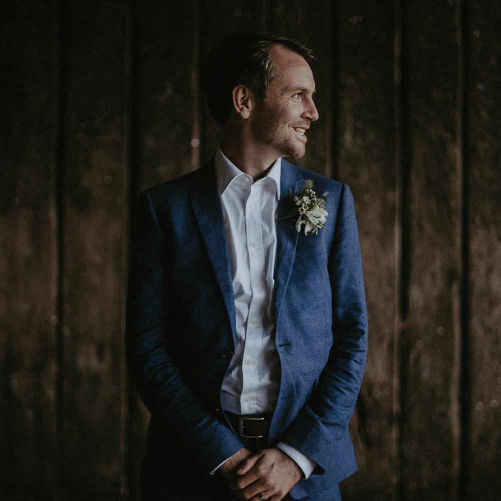 Groom stands with hands crossed and looks to the side smiling on wedding day