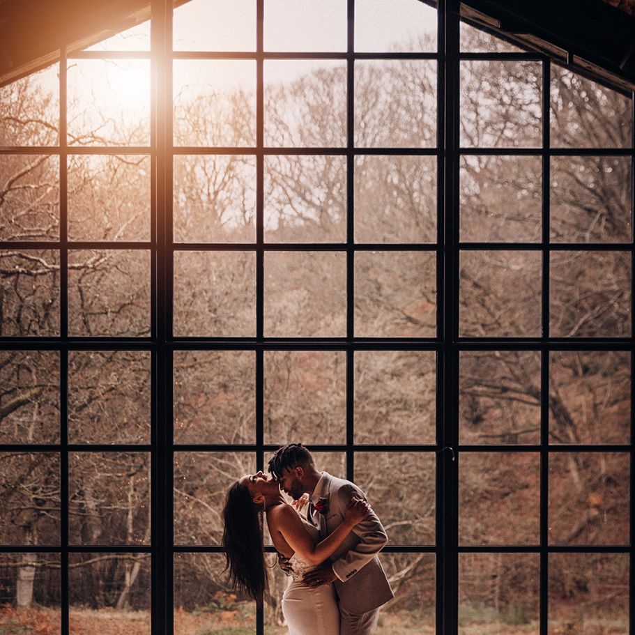 Bride & groom embrace in front of window as the sun sets behind them