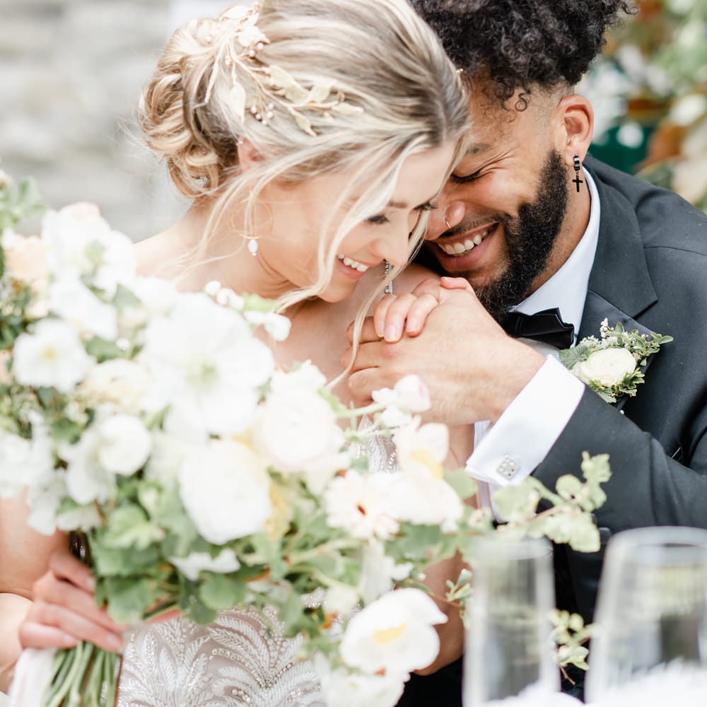 Bearded groom in a black suit embracing his bride at the reception table 
