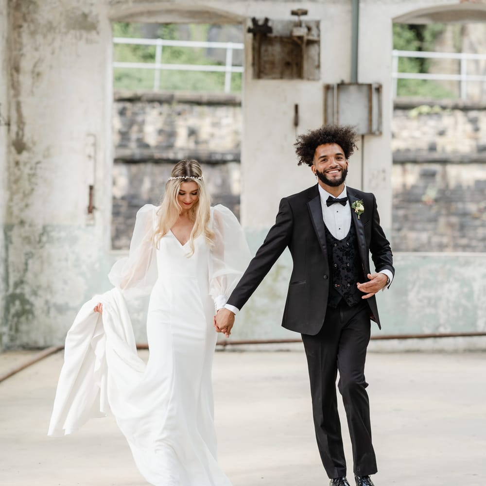 Groom in a three piece black suit with jacquard waistcoat holding hands with his bride in a fitted wedding dress with sheer blouson sleeves 