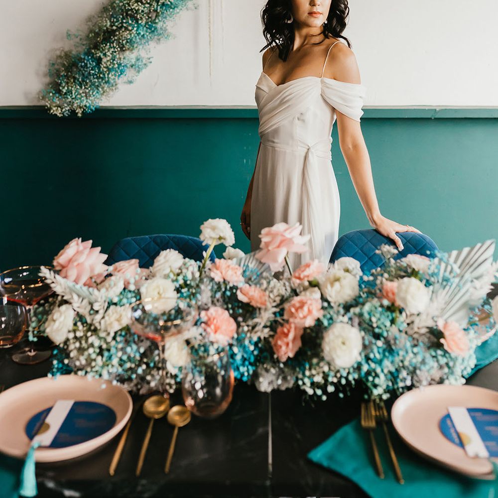Bride in a cold shoulder wedding dress standing next to her reception table decorated in white and teal gypsophila flowers 