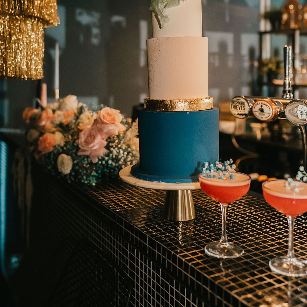 White, light pink, gold and teal wedding cake with sugar shard cake topper resting not the bar with colourful cocktails 