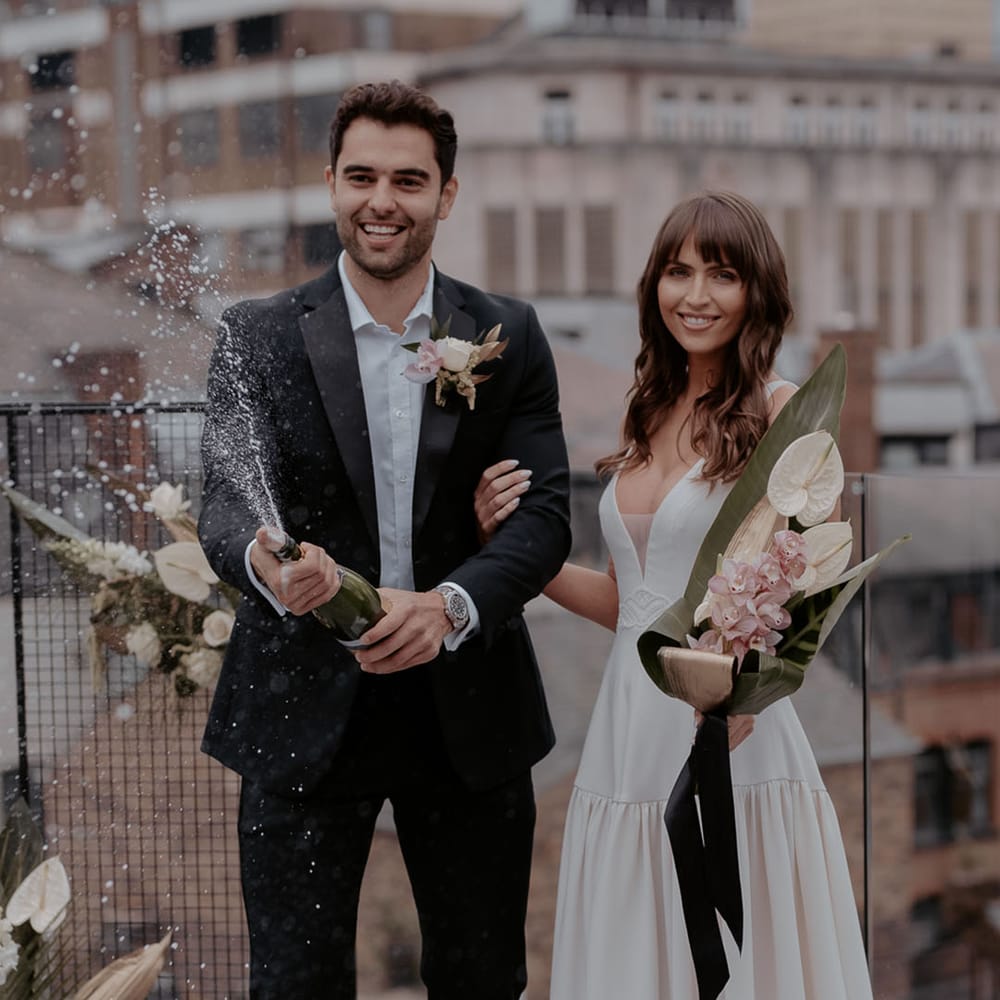 Groom popping champagne by his bride in a Daniel Chu wedding dress holding a anthurium and orchid tropical wedding bouquet 