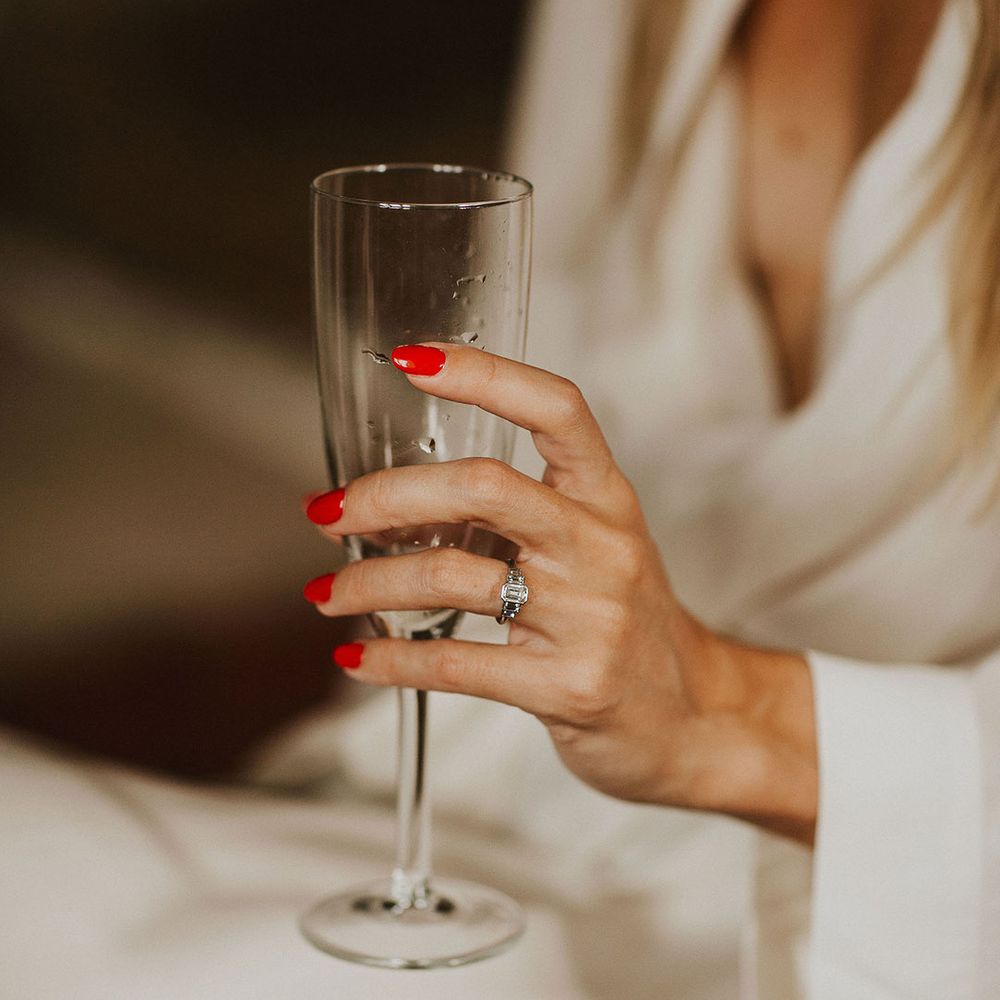Bride holds champagne flute and wears rectangle diamond engagement ring with bright pink nail varnish 