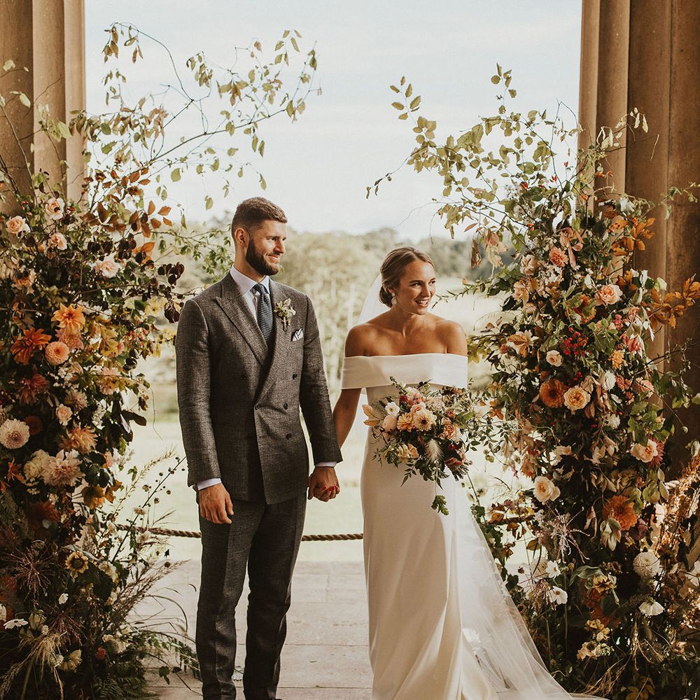 Bride & groom stand under floral archway outdoors in front of large pillars as they hold hands