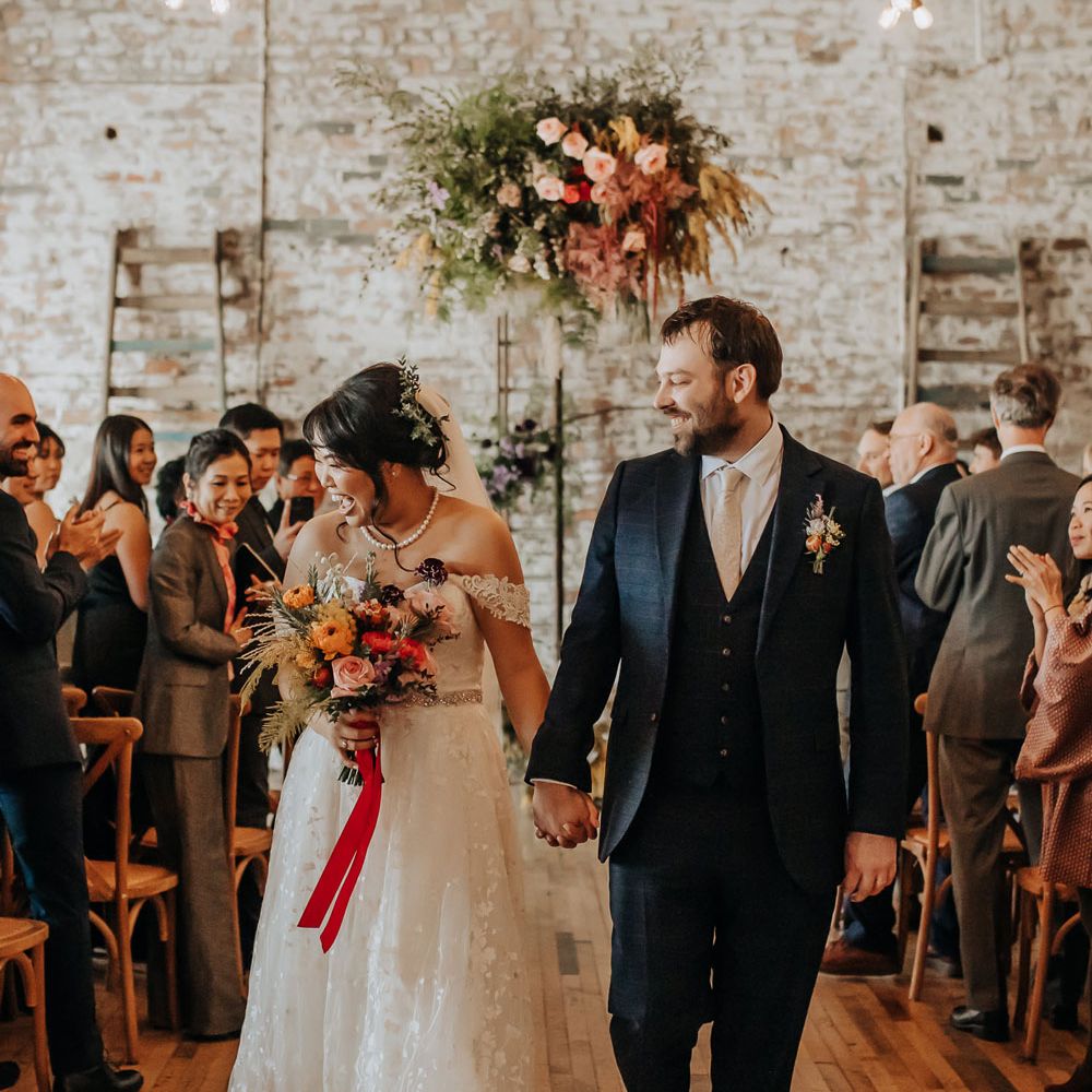 Bride in lace off the shoulder wedding dress and pearl necklace holding orange, pink and green bridal bouquet tied with red ribbon walks down the aisle with groom in dark blue three piece suit
