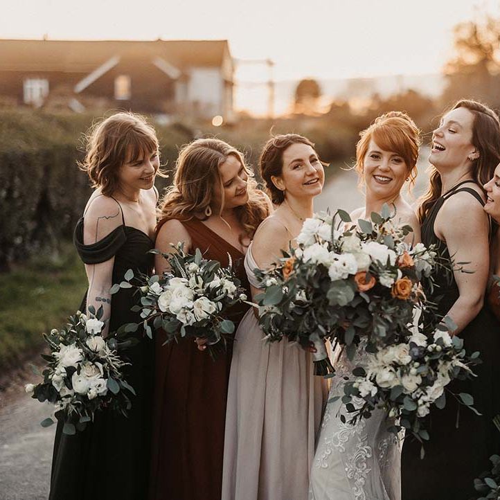 Bride laughs with her bridesmaids in earth toned dresses with various necklines outside barn wedding venue 