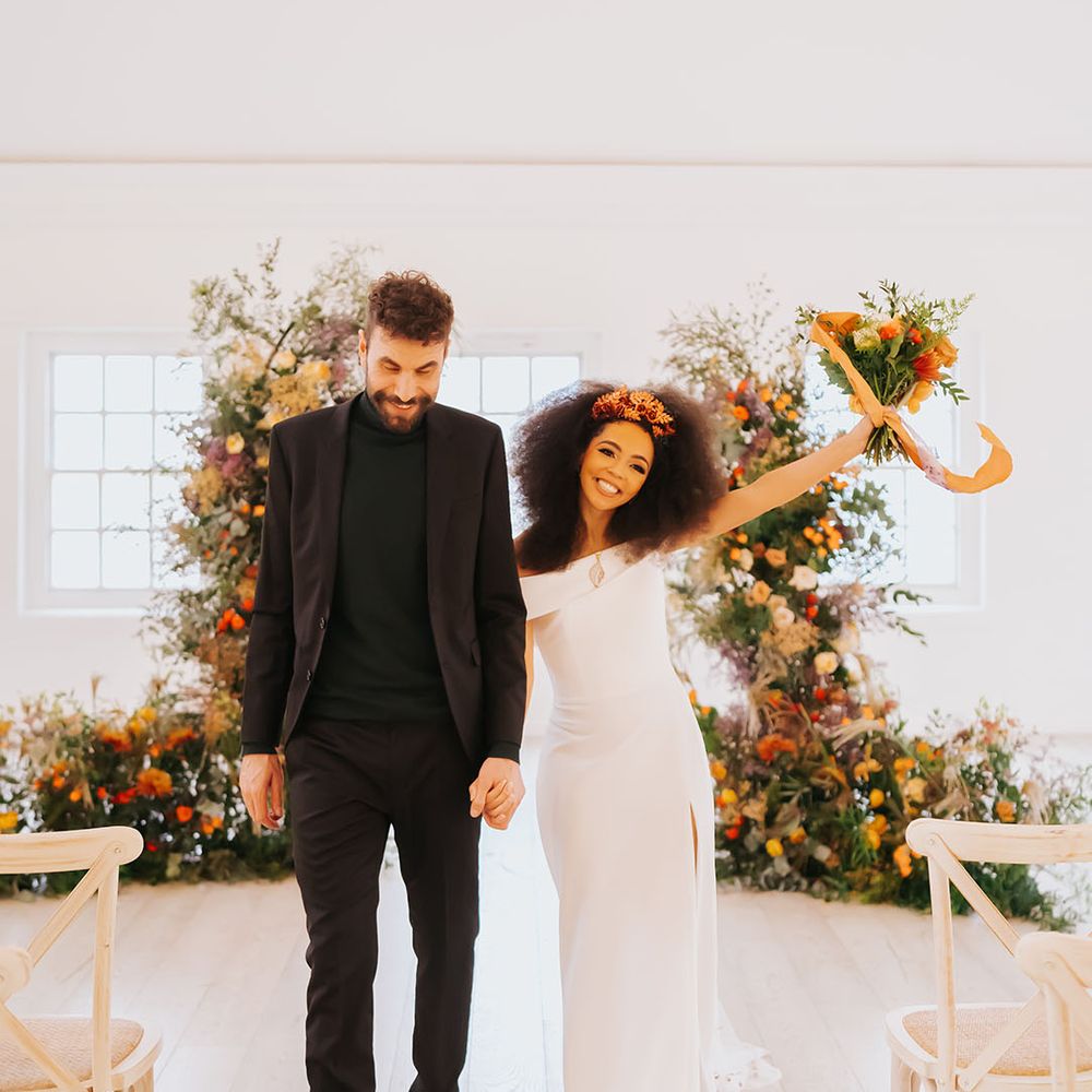 Groom in an all black suit and Black bride with afro hair in an off the shoulder wedding dress with front slit at minimalist golden wedding