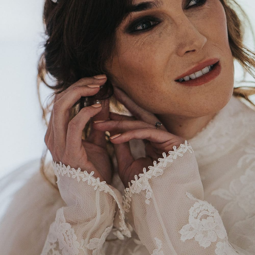 Juno Dawson putting on earrings as she wears long sleeved lace wedding gown with high neck and wedding tiara