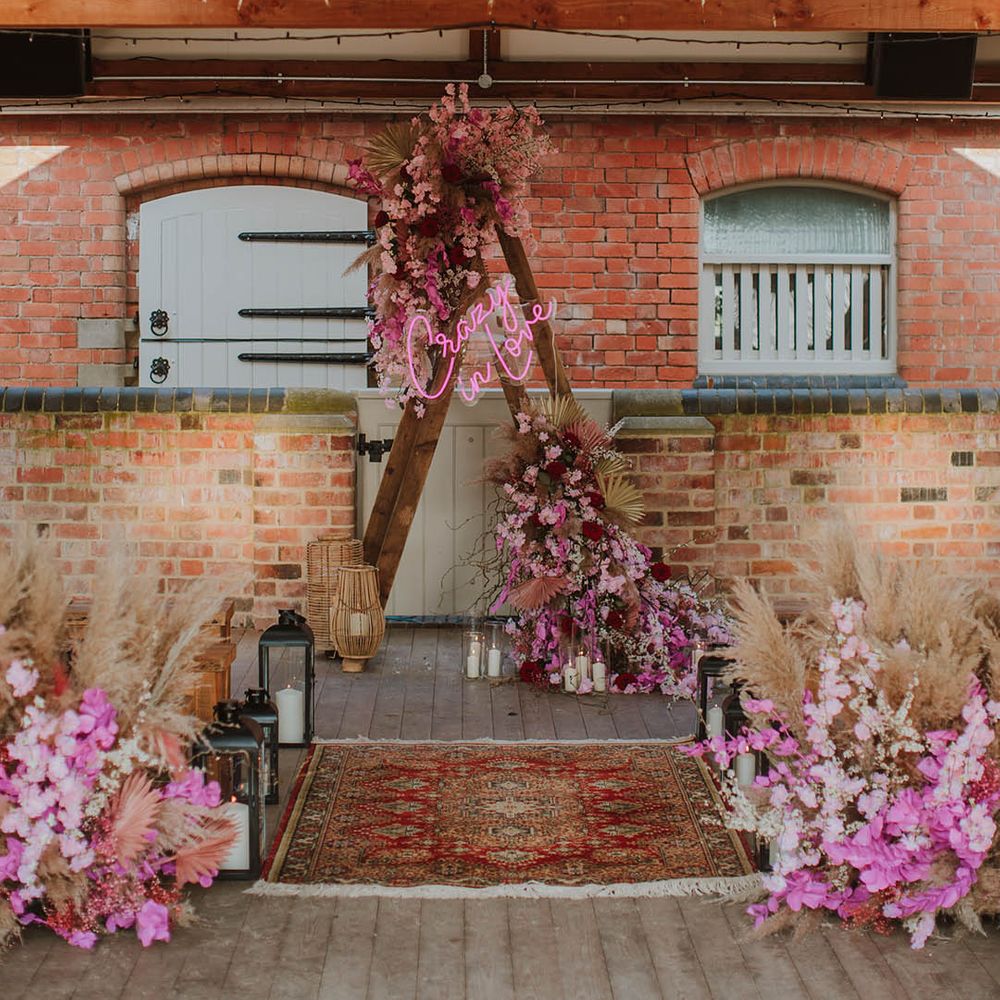 Rustic wedding styling with wooden benches, wool rugs, lanterns and wooden frame altar decorated with a pink neon sign, dried grasses and pink flowers