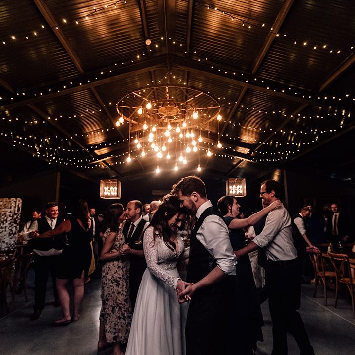 Bride and groom dance together with other wedding guests on the dance floor with chandelier of exposed lightbulbs 
