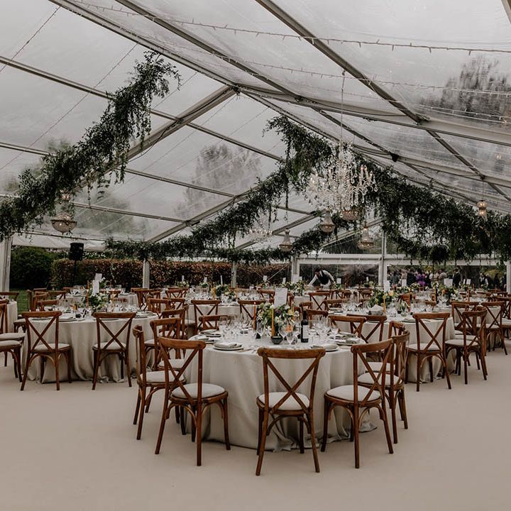 Clear glass marquee wedding reception with wooden furniture and hanging greenery garlands 