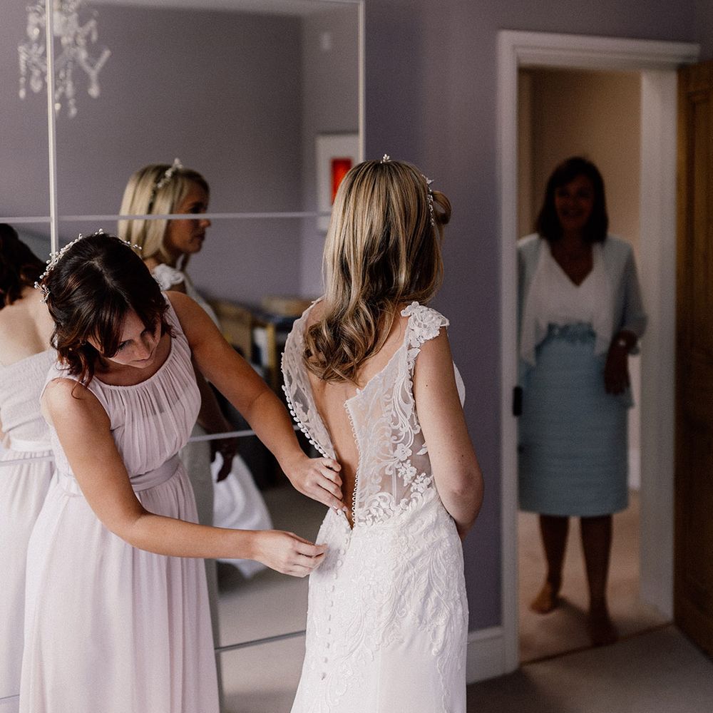 Bridesmaid in blush pink chiffon dress buttons up the bride into her lace wedding dress 