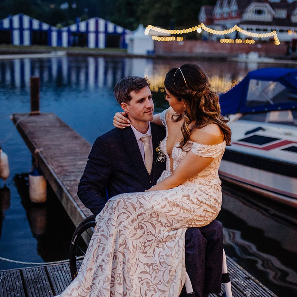 Groom in a blue suit with a gold tie with the bride sitting on his lap wearing an off the shoulder boho lace fitted wedding dress and gold head piece 