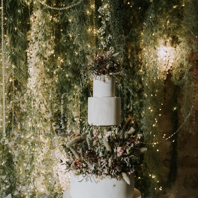 three tier wedding cake with dried and fresh flower layer and cake topper in front of a foliage and fairy light back drop