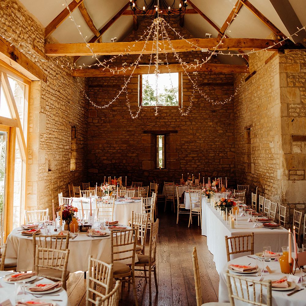 The Barn At Upcote wedding reception finished with round tables complete with white tablecloths, shades of pink and coral decor and hanging fairy lights