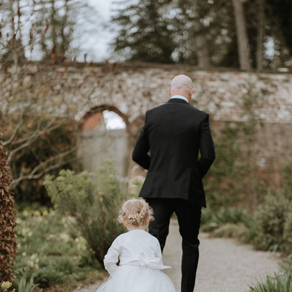 Flower girl in a white dress with a white cardigan and braided updo walks behind the groom 