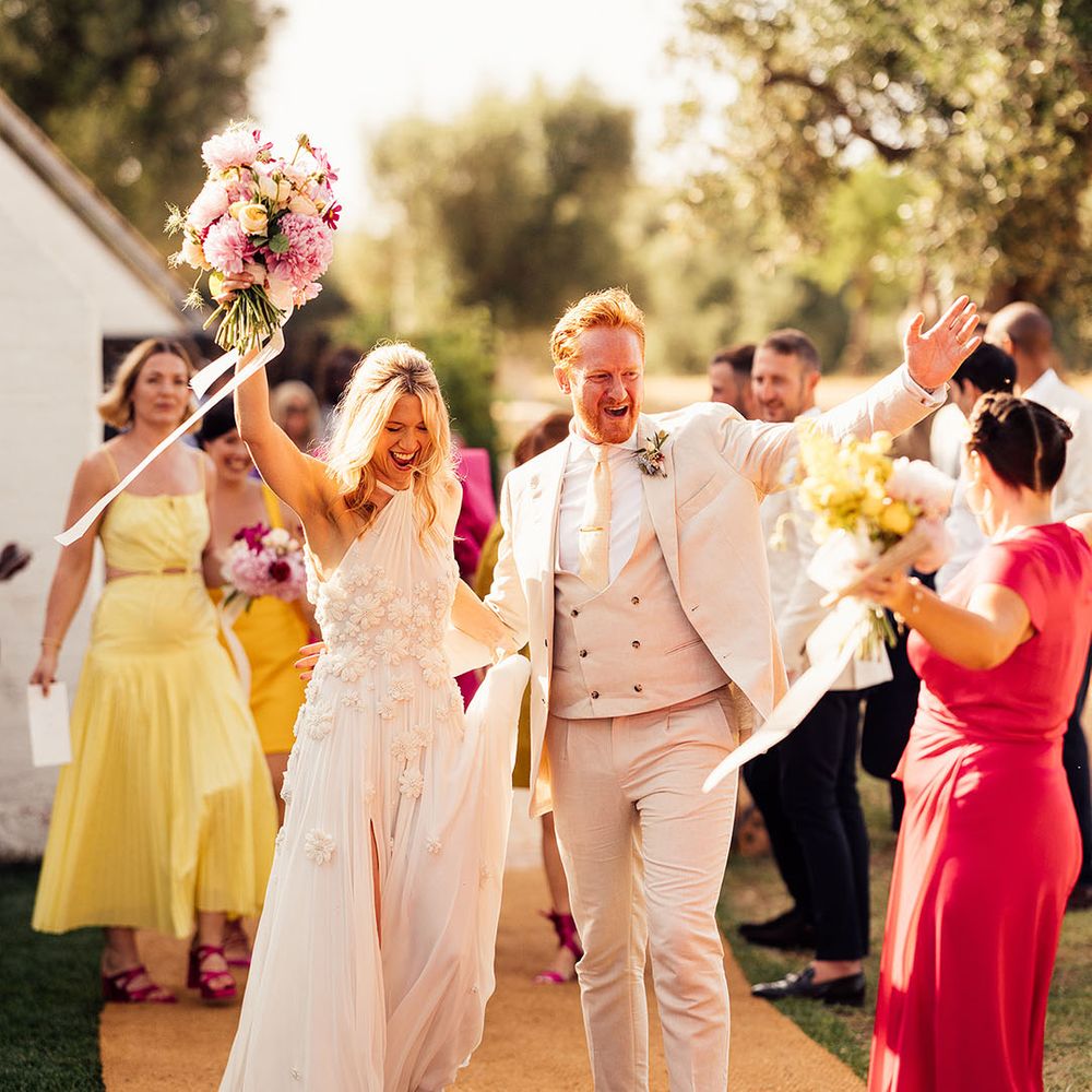 Groom in a cream suit walking with the bride in a halter flower Emma Beaumont wedding dress 