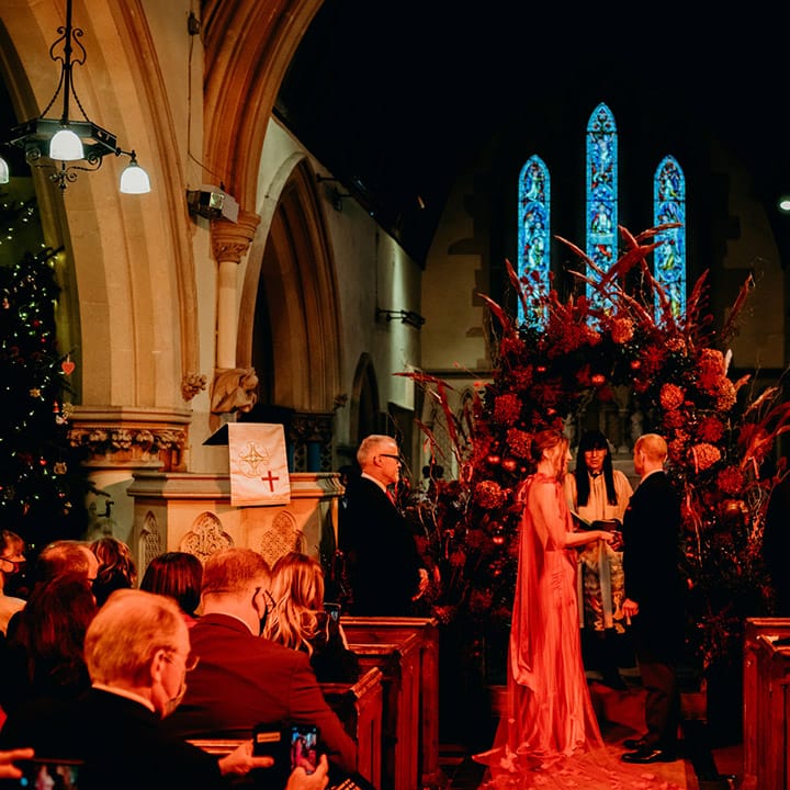 Bride in red satin wedding dress with cape and groom in three piece suit with red tie holding hands at the church alter in front of large Christmas themed floral arch