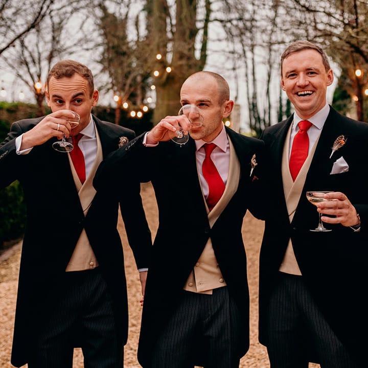 Grooms party hugging and drinking, wearing matching three piece suits, red ties and boutonnieres