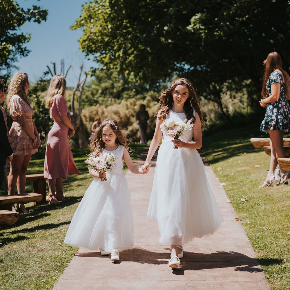 Flower girls walk down the aisle in white dresses with princess skirts holding small floral bouquets 