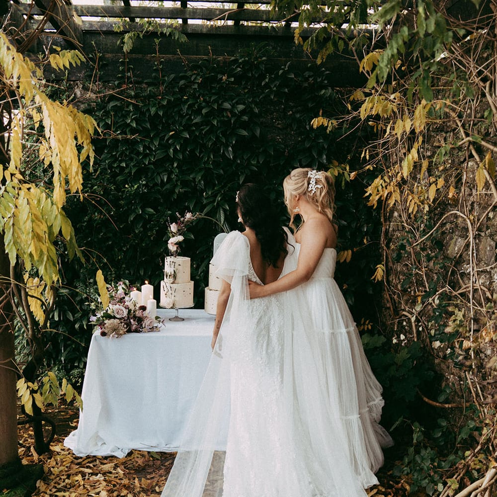 Bride in open back detail lace wedding dress with short puff lace sleeves and bride in sleeveless layered tulle wedding dress at the alter at Holesfoot venue