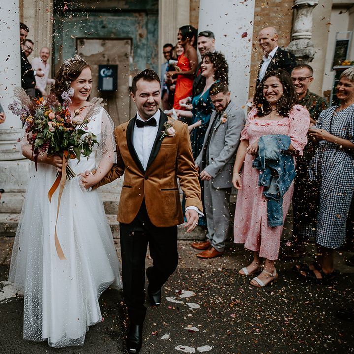 Bride and Groom walk through confetti throw wearing a coloured suit jacket and celestial wedding dress whilst carrying an autumnal bouquet full of Thistles, dried grass and Lila Delphiniums