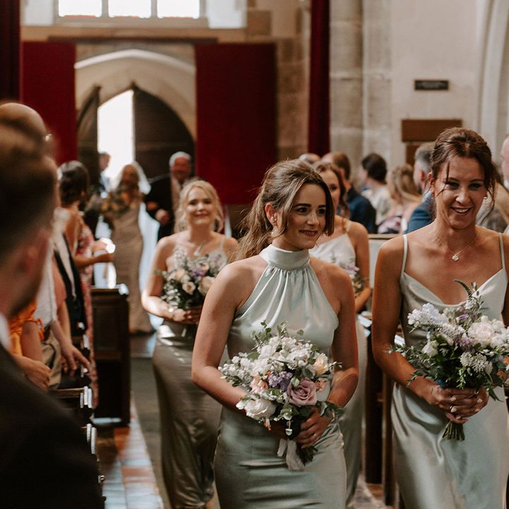 Bridesmaids in sage green satin dresses with different styles walking down the aisle holding small pastel bouquets 