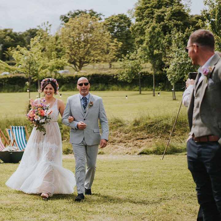 Bride wears colourful flower crown and carries matching bouquet as she walks down the aisle with her father outdoors 