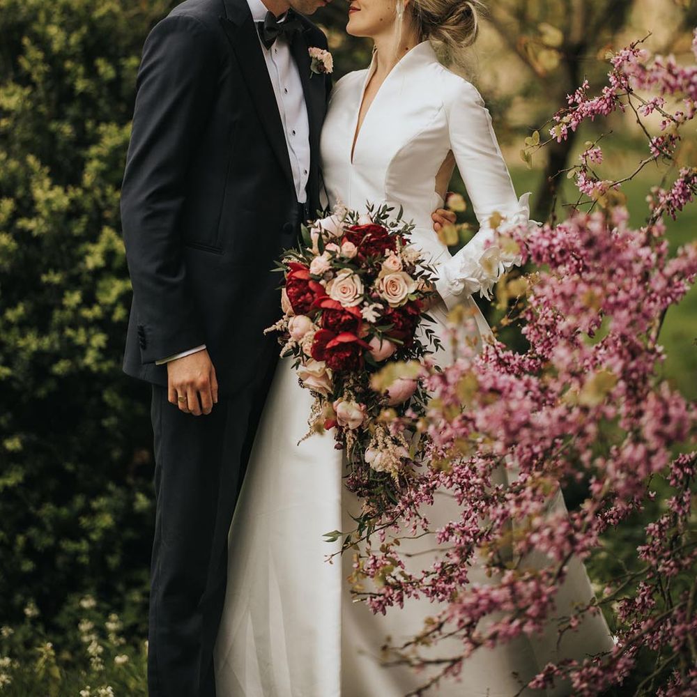The groom in black tie with a white rose buttonhole leans in to kiss the bride in a long sleeve high neck Jesus Peiro wedding dress with floral hair piece