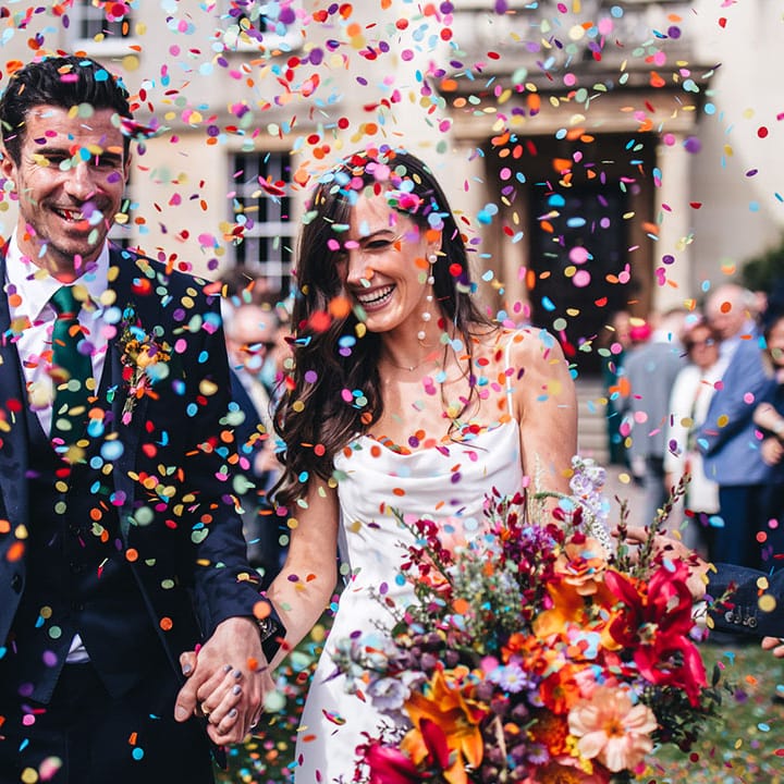 Groom in three piece navy suit with green tie standing with bride in satin wedding gown walking out to colourful confetti 