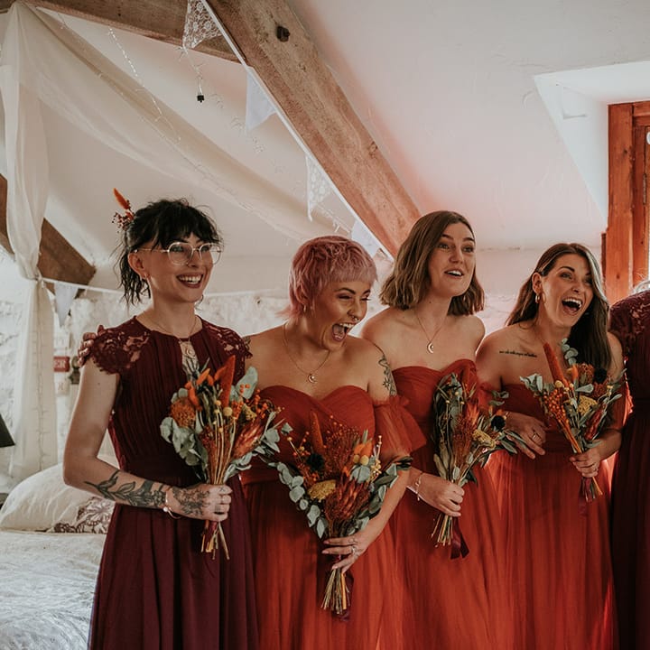 Mismatched burgundy and burnt orange bridesmaid dresses holding dried flower and eucalyptus bouquets doing first look with bride 