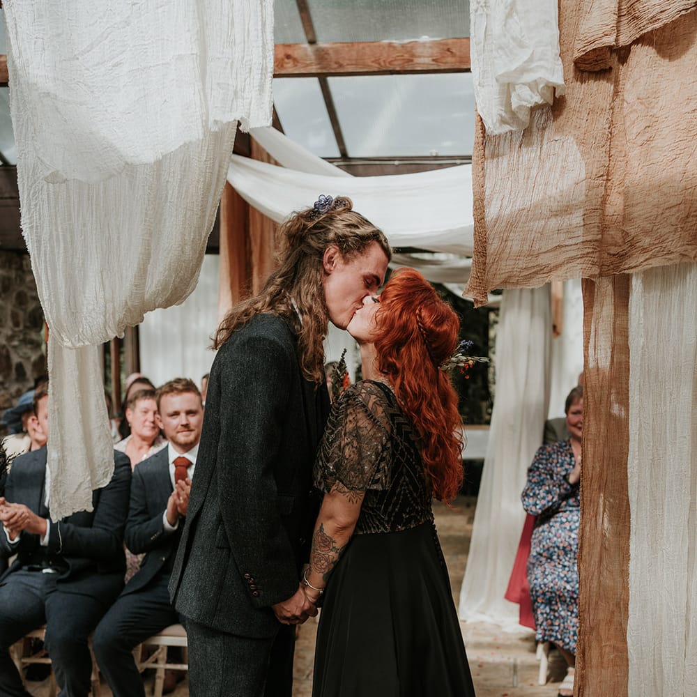 Groom in dark grey three piece suit and burgundy flower patterned tie kissing bride in embellished black wedding dress with sheer sleeves standing in Westcott Barton wedding venue with off white and orange ceiling drapes 