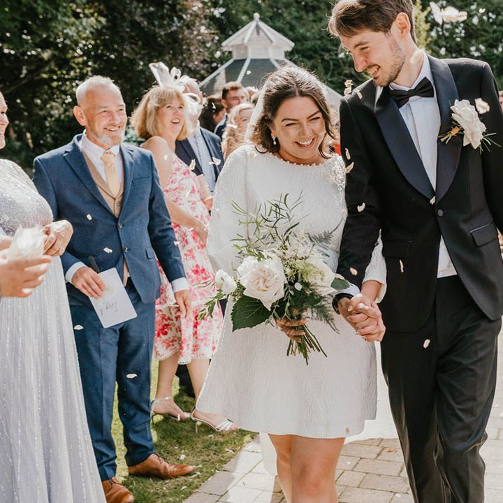 The bride wears a high neck long sleeve short wedding dress walking with groom in black tie for their confetti exit 
