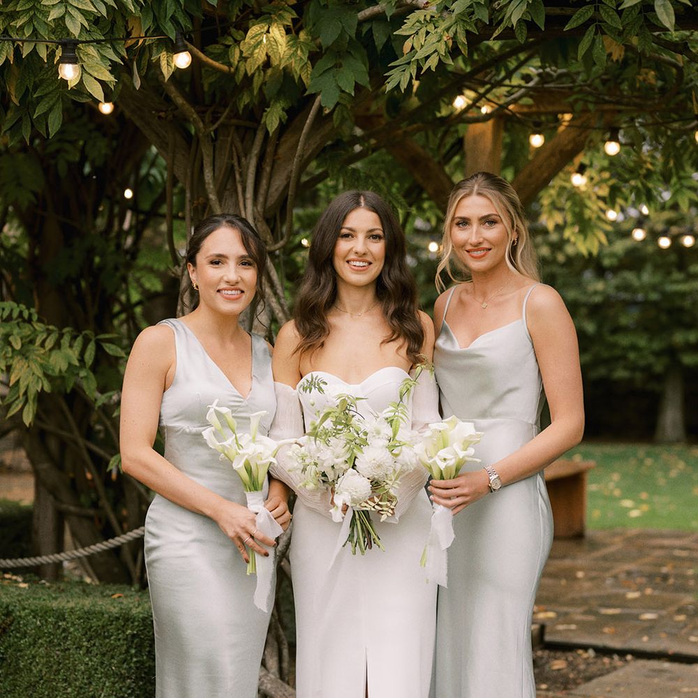 Bridesmaids in mismatched sage green dresses with the bride in a strapless corset wedding dress alll holding with flower bouquets with lilies, dahlias and more