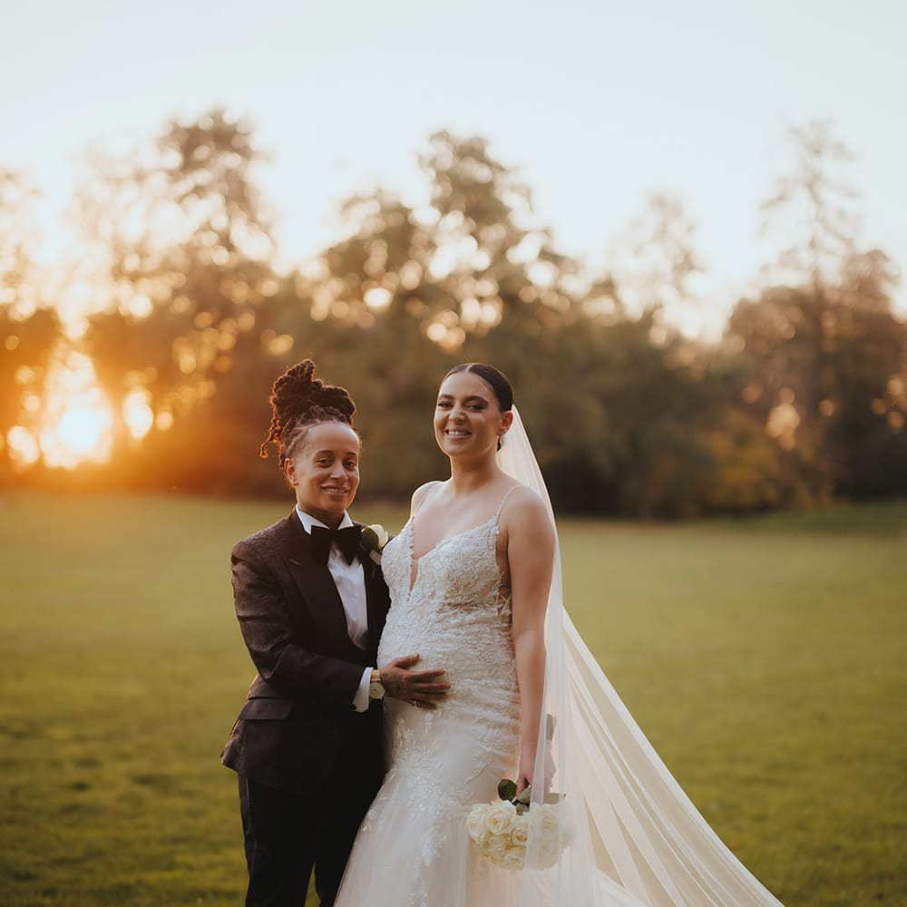 Bride in lace sleeveless v neck Enzoani wedding dress with tulle finish and church length veil posing with bride in black snakeskin print tuxedo with black bowtie and white rose boutonniere at golden hour at Grittleton House lesbian black tie wedding 