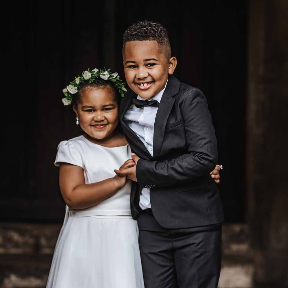 Flower girl wearing short sleeve satin dress and flower crown standing with page boy in black suit and bowtie 