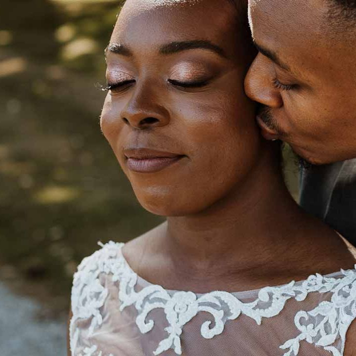Bride wearing lace and mesh boatneck wedding dress smiling with groom in charcoal grey grooms suit, light grey patterned tie and large white rose and foliage boutonniere
