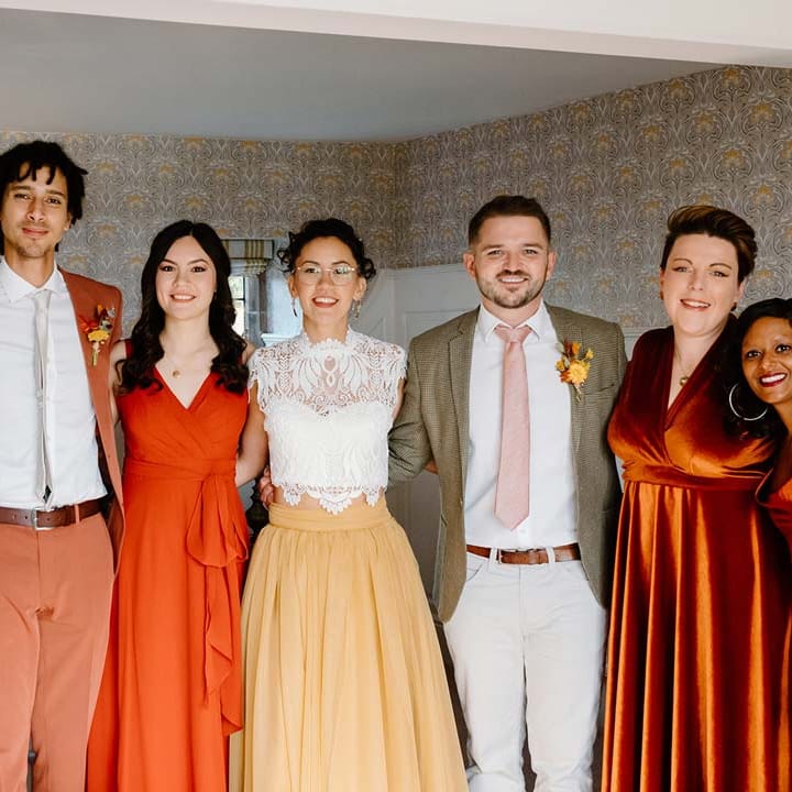 Bride wearing white mesh lace bridal crop top and yellow wedding skirt standing with bridesmaids in mismatched bridesmaid dresses and wedding guests in yellow and orange wedding theme