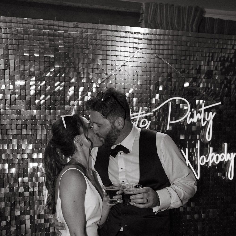 Bride and groom share a kiss with there cocktails in front of neon wedding sign and glitter wall 